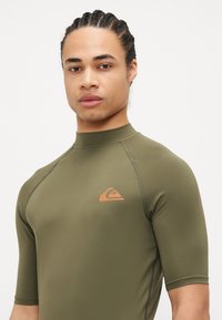 Olivovo zelený rash guard s krátkym rukávom vyrobený z elastického materiálu so plochými švami. Obsahuje malé oranžové logo na hrudi.