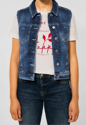 Vrouw met een blauw spijkervestje over een wit T-shirt met rode tekst en donkerblauwe jeans, staand tegen een effen achtergrond.