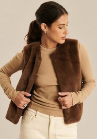 Brązowa kamizelka z sztucznego futra bez rękawów, noszona na beżowej bluzce z długim rękawem. Gładka tekstura i otwarty przód, zestawiona z jasnymi dżinsami.