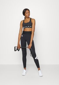 Sustentação desportiva preta com detalhes brancos no logótipo e leggings altas pretas com um desenho branco. Modelo segura bandas de exercício, usando ténis brancos.