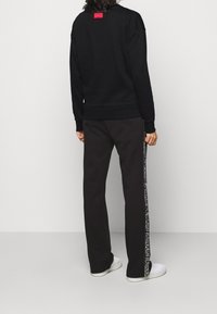 Persona che indossa un felpa nera con il logo rosso di Hugo sulla schiena, pantaloni neri con strisce laterali bianche e sneakers bianche, in piedi su un pavimento grigio.