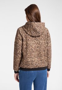 Donna che indossa una giacca con cappuccio a stampa leopardata marrone e nera con jeans blu, vista di spalle su sfondo bianco uniforme.