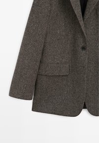 Blazer en laine tachetée marron foncé avec un bouton noir visible et une poche à rabat sur le devant sur fond blanc.