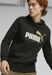 Sudadera negra con bolsillo canguro, presenta un gran logotipo de "PUMA" en blanco y dorado, con "1948-2023-75" en verde debajo. Tejido suave.