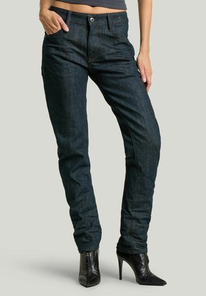 Femme portant un jean slim bleu foncé et des bottines noires à talons hauts, une main dans la poche, debout sur un fond neutre.
