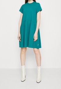 Tyrkysové bavlněné šaty s krátkým rukávem, se sbíraným pasem, rozšířenou sukní a kulatým výstřihem. Doplňují je bílé kotníkové boty.