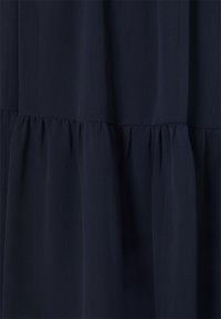 Tummansininen kangas, jossa on kerätty röyhelöyksityiskohta, luomassa kerroksellista vaikutusta vaatteeseen.