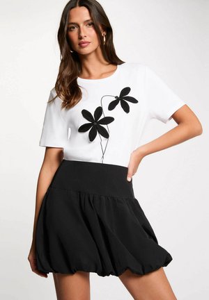 Femme aux longs cheveux bruns portant un haut blanc avec un motif de fleur noire et une jupe noire taille haute avec un ourlet froncé.