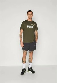 Férfi olajzöld póló fehér "PUMA" logóval, fekete rövidnadrággal és fekete, lime zöld kiegészítőkkel ellátott sportcipővel párosítva, semleges háttér előtt bemutatva.