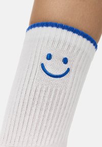 Hvide rillede sokker med en blå topkant og en broderet blå smiley ansigt på benet. Blød tekstur, designet til komfort og afslappet brug.