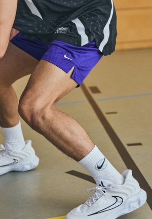 Person trägt lila Nike-Shorts, weiße Nike-Socken und weiße Nike-Sneakers auf einem Hallen-Basketballplatz.