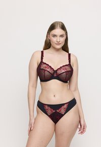 Kobieta z długimi jasnobrązowymi włosami, ubrana w czarny i czerwony koronkowy stanik w kwiaty oraz pasujące majtki, stojąca na jasnym tle.