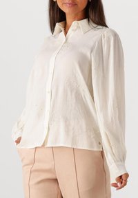 Vrouw in een wit blouse met lange mouwen en subtiele geborduurde bloemenpatronen, gecombineerd met beige hoge taillebroek.