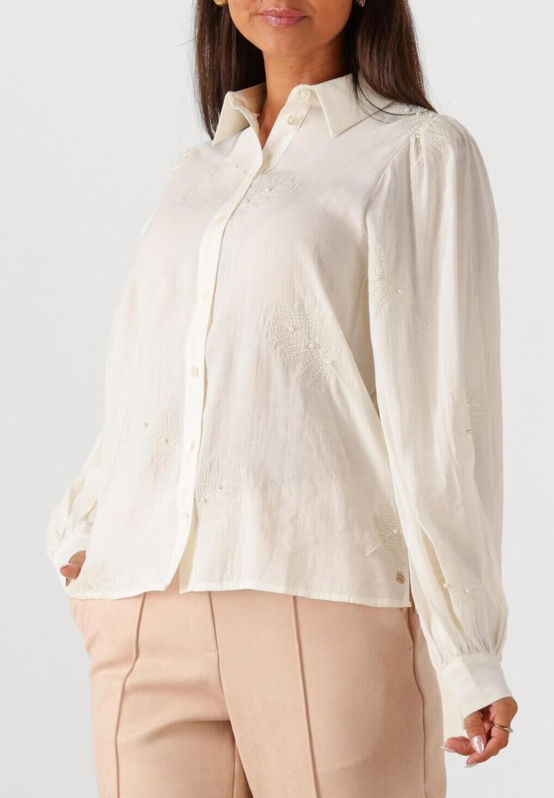 Vrouw in een wit blouse met lange mouwen en subtiele geborduurde bloemenpatronen, gecombineerd met beige hoge taillebroek.