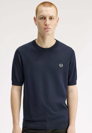 Jonge man met kort rood haar die een marineblauw kortemouwentrui draagt met een klein wit lauwerkranslogo op de borst.