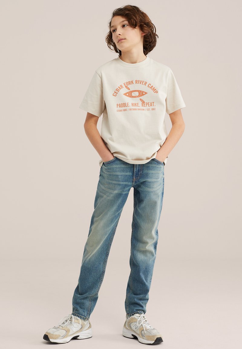 Mladić stoji s rukama u džepovima, nosi kremastu majicu s grafičkim motivom, plave traperice i bijele tenisice na jednobojnoj pozadini.