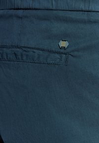 Donkerblauwe stoffen shorts met een gladde textuur, voorzien van een hexagonaal metalen accent en een klein zakje op de achterkant.