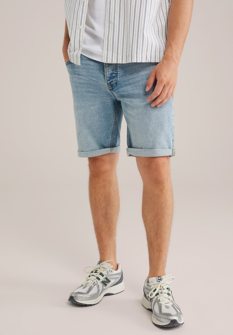 Moški v svetlo modrih zavihanih denim kratkih hlačah, belih in srebrnih supergah, beli majici ter lahki črtasti majici s kratkimi rokavi, ki ima roke v žepih.