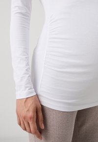 White long-sleeve top with gathered side seams. Pair of beige ribbed pants visible below. Hand resting on hip shows natural skin tone.