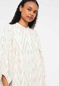 Vestido branco com padrão de ziguezague dourado, tecido texturizado, decote alto e mangas bufantes. Silhueta fluida com detalhe de cintura franzida.