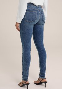 Jeans de mezclilla en azul medio con un corte ajustado, cintura alta y dos bolsillos traseros. Combinados con tacones de tiras negros. Textura suave y ligeramente desgastada.