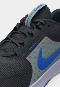 Αθλητικό παπούτσι σε μαύρο και γκρι χρώμα με μπλε λογότυπο Nike, γκρι κορδόνια και θηλιά στη γλώσσα με την επιγραφή "NIKE SP RUNNING S" και "DYNAMIC FIT" στο πλάι.