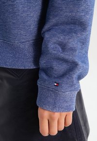 Sudadera azul con un acabado texturizado, que presenta puños acanalados y una pequeña etiqueta de logo en rojo, blanco y azul cerca de la muñeca. Falda negra visible.