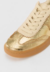 Gold leather sneaker with a textured finish, cream laces, scalloped detailing, and a gum rubber sole.