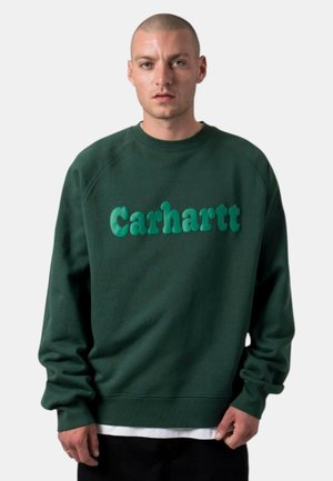 Uomo che indossa una felpa Carhartt verde scuro con logo in rilievo, pantaloni neri e maglietta bianca sotto, in piedi davanti a uno sfondo chiaro e uniforme.