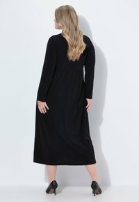 Kobieta z długimi blond włosami ubrana w czarną midi sukienkę z długim rękawem i czarne szpilki, stoi przodem do białej ściany z cieniami za sobą.
