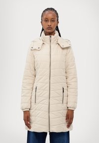 Manteau long beige matelassé avec col montant, fermeture éclair et deux poches latérales. Présente un motif texturé et des pressions aux épaules pour ajustement.