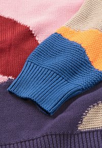 Pull en tricot coloré avec une manche texturée bleue, comprenant des sections en rose, rouge, orange et violet, et des poignets côtelés.