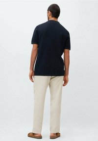 Korte mouwen, marineblauwe polo uit gestructureerde stof; gecombineerd met lichtbeige broeken en bruine instappers. Neutrale achtergrond.