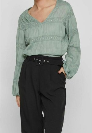 Femme portant un chemisier vert clair texturé avec des détails en dentelle et un pantalon noir taille haute avec une ceinture noire, main dans la poche.