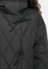 Μαύρο επενδυμένο μπουφάν με ψηλό γιακά, που διαθέτει κλείσιμο με κορδόνια στο μπροστινό μέρος. Το ύφασμα έχει λεία υφή και διακοσμείται με σχέδιο διαμαντιού.