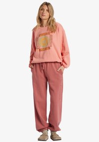 Sweatshirt rose pastel avec un graphisme de soleil, associé à un pantalon rose ample. Les deux articles ont une texture douce, avec un subtil détail de broderie sur le pantalon.