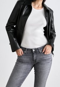 Chaqueta negra de cuero sintético con cremallera frontal, camiseta blanca de canalé y vaqueros ajustados grises con detalles de botones y bolsillos.