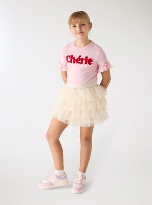Młoda dziewczyna stoi z rękami za plecami, ubrana w różowy top "Chérie", warstwową kremową spódnicę tutu, białe skarpetki i różowo-białe trampki.
