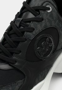 Μαύρο αθλητικό παπούτσι με υφή σε γκρι λεπτομέρειες, στρογγυλό λογότυπο και αντίθετες λείες επιφάνειες. Οι κορδόνια είναι μαύρα, με επένδυση στον αστράγαλο για άνεση.