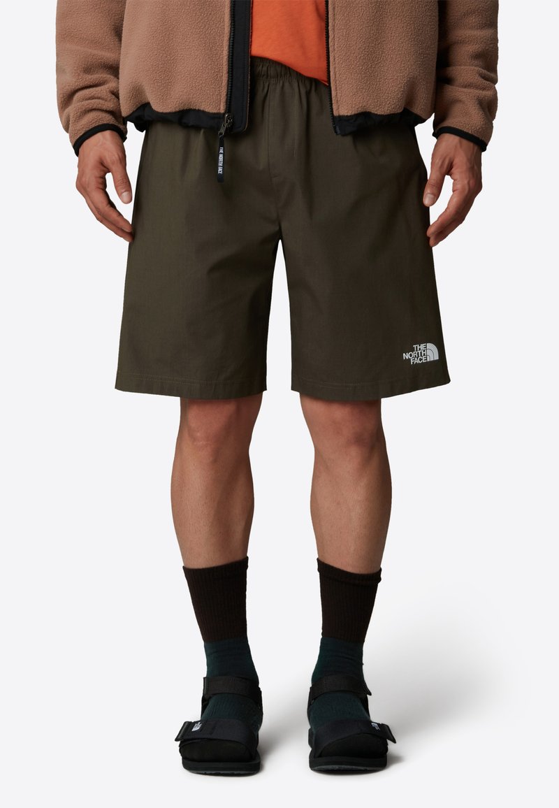 Shorts vert olive avec une taille élastique, un logo blanc sur la jambe gauche, associés à des sandales noires et des chaussettes de crew marron foncé.