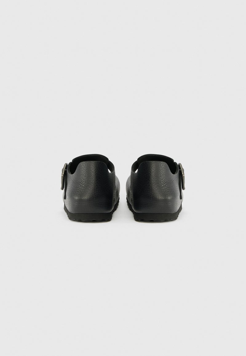 Schwarze Hinteransicht von Schuhwerk mit strukturierter Oberfläche, offenem Absatz und seitlichem Schnallendetail; flache Gummisohle mit Rillenmuster.