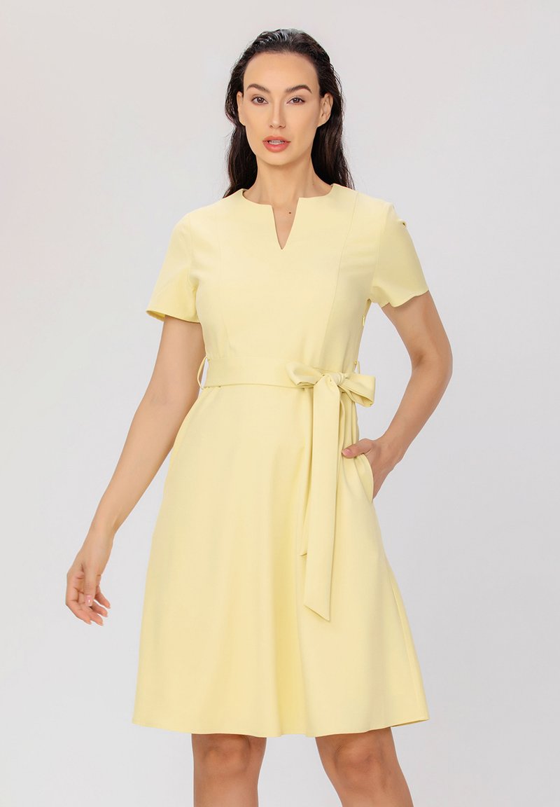Robe jaune à mi-mollet avec des manches courtes, un décolleté en V et une ceinture nouée à la taille, comportant des poches sur les côtés et un tissu lisse.