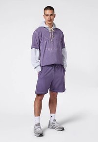 Sudadera de algodón morada con capucha y cordones, sobre una camiseta gris de manga larga, combinada con pantalones cortos morados a juego y zapatillas deportivas grises.