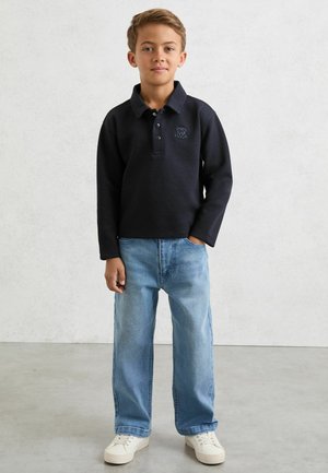 Námořnicky modrá polokošile s dlouhým rukávem a vyšitým logem, doplněná světle modrými džínami s širokými nohavicemi a bílými teniskami, pózující na neutrálním pozadí.