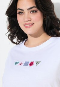 T-shirt en coton blanc avec des formes brodées colorées et scintillantes : cœur, étoile, carré, cercle et triangle, dans des couleurs et motifs variés.
