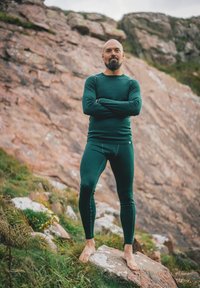 Muž stojící bos na kameni se založenýma rukama, oblečený v tmavě zeleném termoprádle, na pozadí skalnatého svahu a trávy.