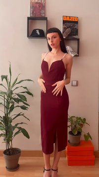 Γυναίκα με αμάνικο μπορντό φόρεμα με ιμάντες από πέρλες στέκεται μέσα σε εσωτερικό χώρο κοντά σε φυτά, πορτοκαλί κουτιά και ραφιέρες τοίχου που κρατούν περιοδικά και καπέλα.