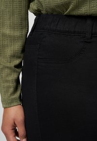 Jupe en denim stretch noire avec une taille élastique, des poches sur les côtés et un haut vert olive texturé visible sur le côté gauche.