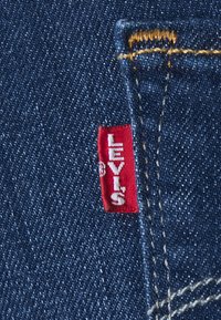 Tecido de denim azul-escuro com um rótulo vermelho da Levi's preso. A costura é dourada, destacando os detalhes da costura do bolso.