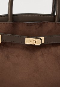 Sac à main en daim marron avec des poignées en cuir lisse, une fermeture à boucle métallique et des détails de couture subtils sur la bandoulière.